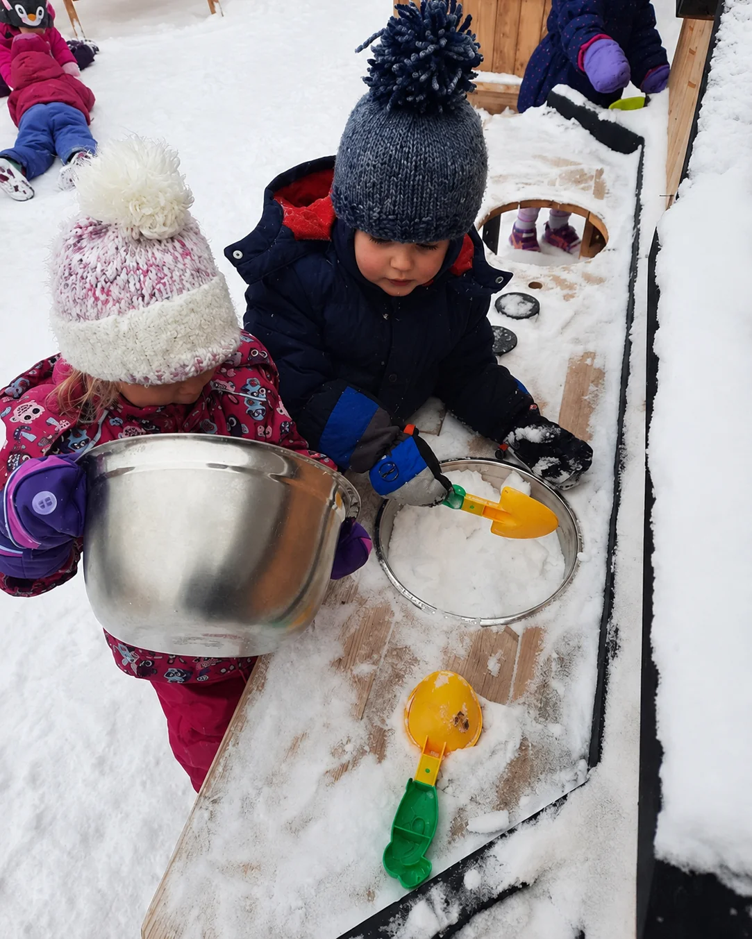 Two children exploring snow with a metal bowl, pan and scoops during outdoor learning.