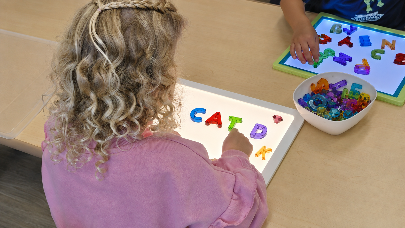 Children using translucent alphabet letters to build words on illuminated classroom light boards.